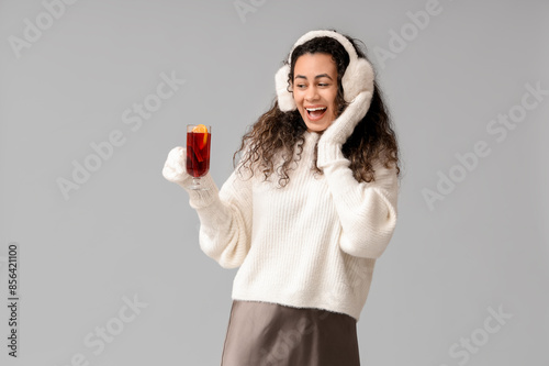 Beautiful young African-American woman in warm mittens with glass cup of hot mulled wine on grey background