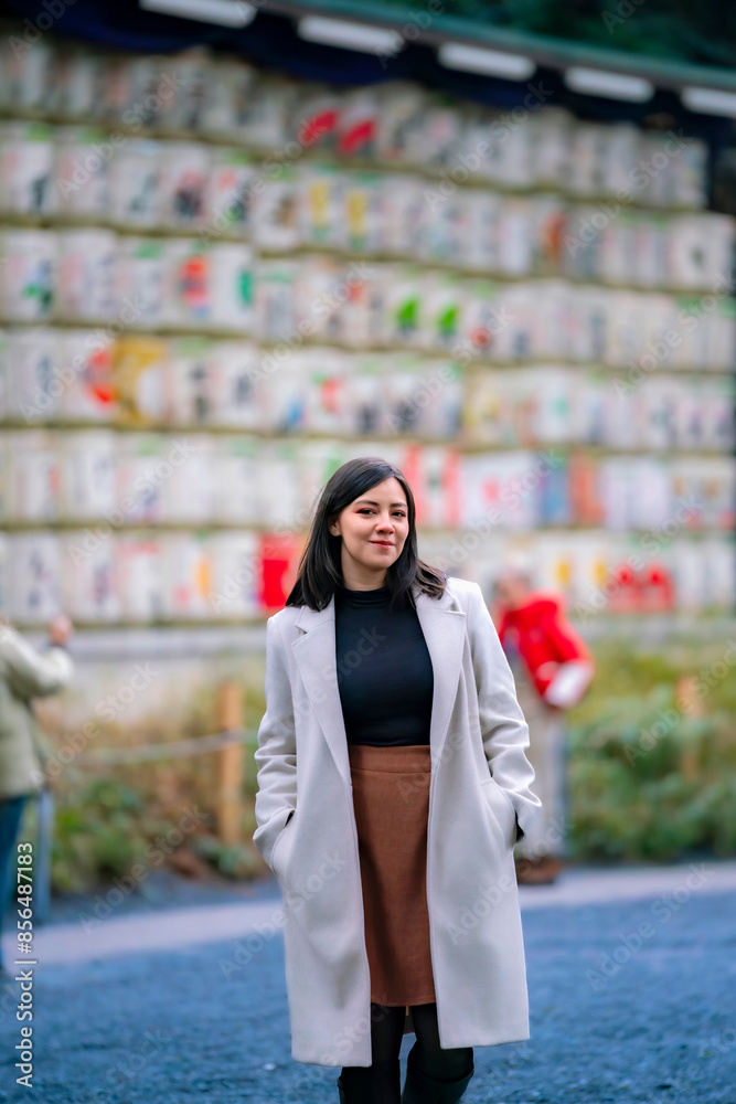 Fototapeta premium A young Latina woman standing in front of decorative sake barrels at Meiji Shrine in Tokyo, Japan, looking content.