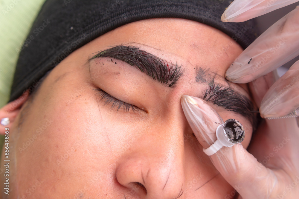 Fototapeta premium An esthetician using a microblade tool to make small cuts into the eyebrow of a customer. A Microblading technique to implant pigment. Male brow enhancement.