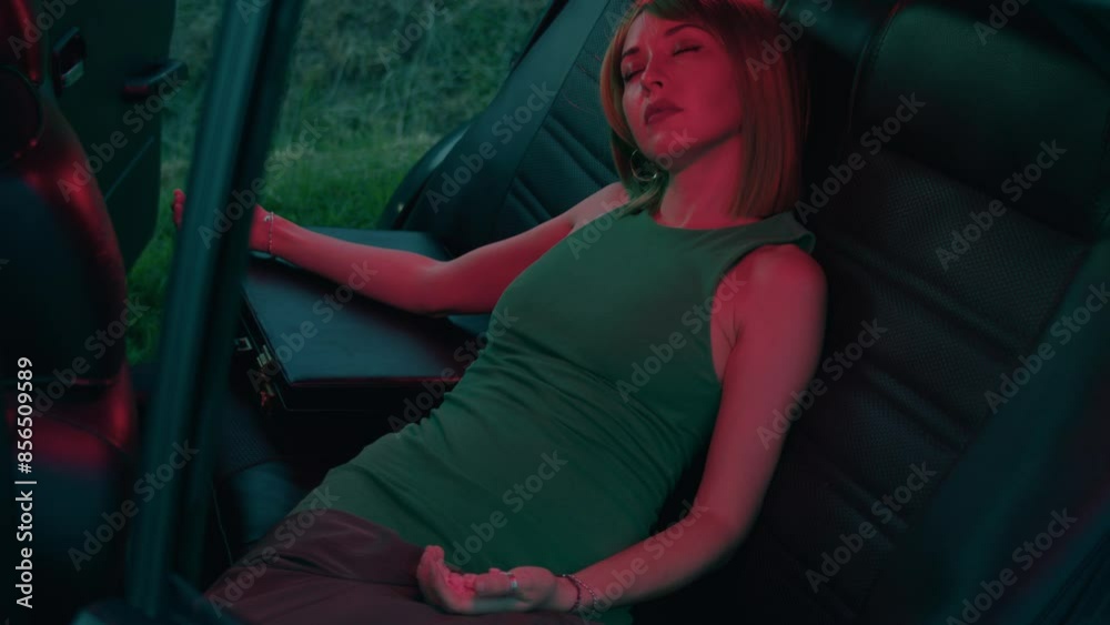 A young stylish woman in a green dress and green wig sits with her eyes closed in a retro car in a red light with flowers all around. A young stylish woman in a green dress and green wig sits with her eyes closed in a retro car in a red light with flowers all around.