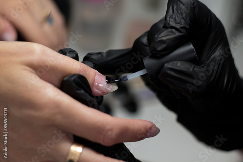 A nail technician wearing black gloves applies nail polish to a client’s nail with precision. The client’s hand is relaxed, highlighting the meticulous attention in the manicure process.