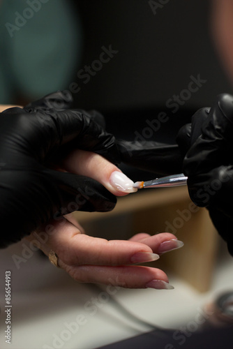 A close-up of a nail technician’s hands in black gloves applying gel polish on a client’s nail. The client’s hand is relaxed, highlighting the precision involved in the nail care process.