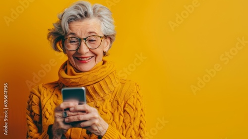 Senior woman with gray hair and glasses smiling while using smartphone. Wearing a mustard yellow sweater and standing against a vibrant yellow background