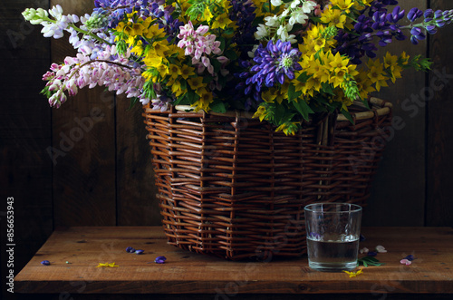 dark rural still life with lupines in a wicker basket and a glass of water. summer rural composition. copy space.