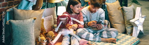 Banner of happy little girl embracing rag doll while looking to boy playing video game with mobile phone sitting on shelter tent at home. Children having fun using cellphone in a cozy diy teepee.