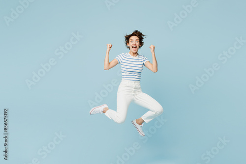 Full body young woman wears striped t-shirt casual clothes jump high doing winner gesture celebrate clenching fists isolated on plain pastel light blue background studio portrait. Lifestyle concept.
