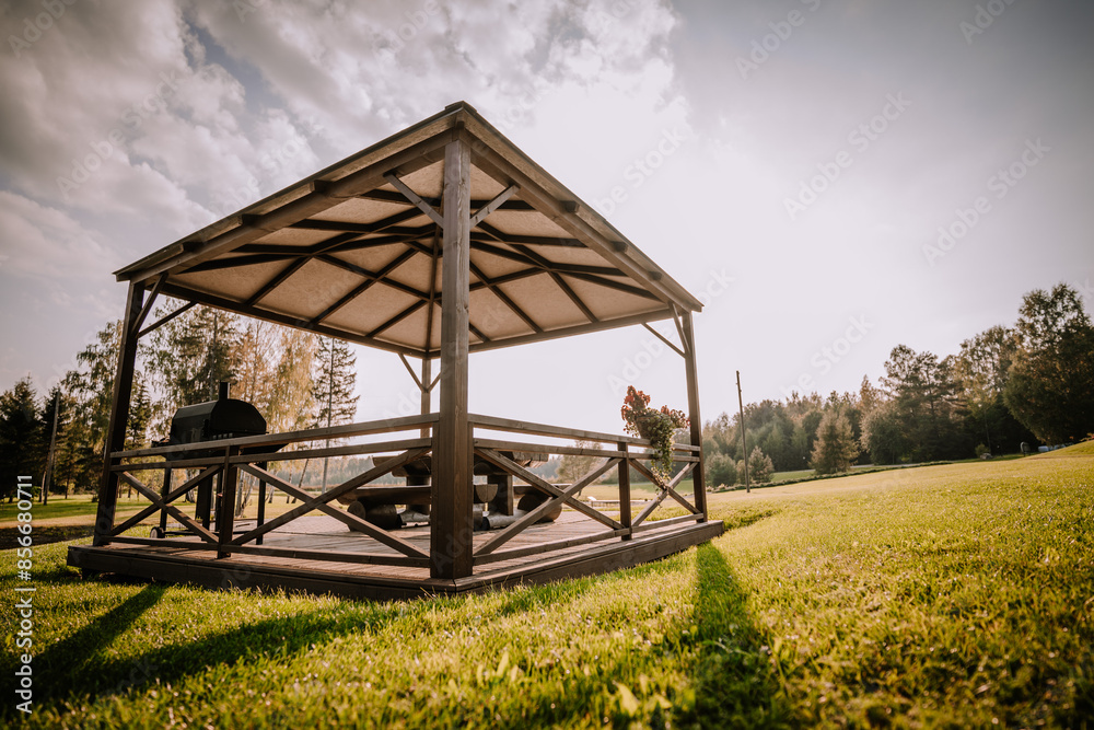 Blome, Latvia - September 11, 2023 - Low-angle view of a wooden gazebo with a grill and picnic table, set in a lush green field under a partly cloudy sky, creating a serene outdoor space. Blome, Latvia - September 11, 2023 - Low-angle view of a wooden gazebo with a grill and picnic table, set in a lush green field under a partly cloudy sky, creating a serene outdoor space.
