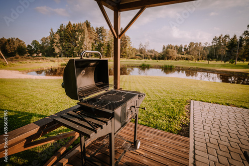 Blome, Latvia - September 11, 2023 - An outdoor grill with an open lid on a wooden deck, overlooking a serene pond and grassy landscape in a park setting. photo