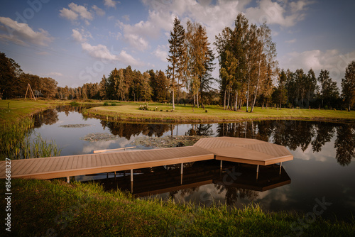 Blome, Latvia - September 11, 2023 - Tranquil lakeside scene with a wooden dock, surrounded by lush trees and greenery, reflecting on the calm water under a blue sky. Copy space. photo