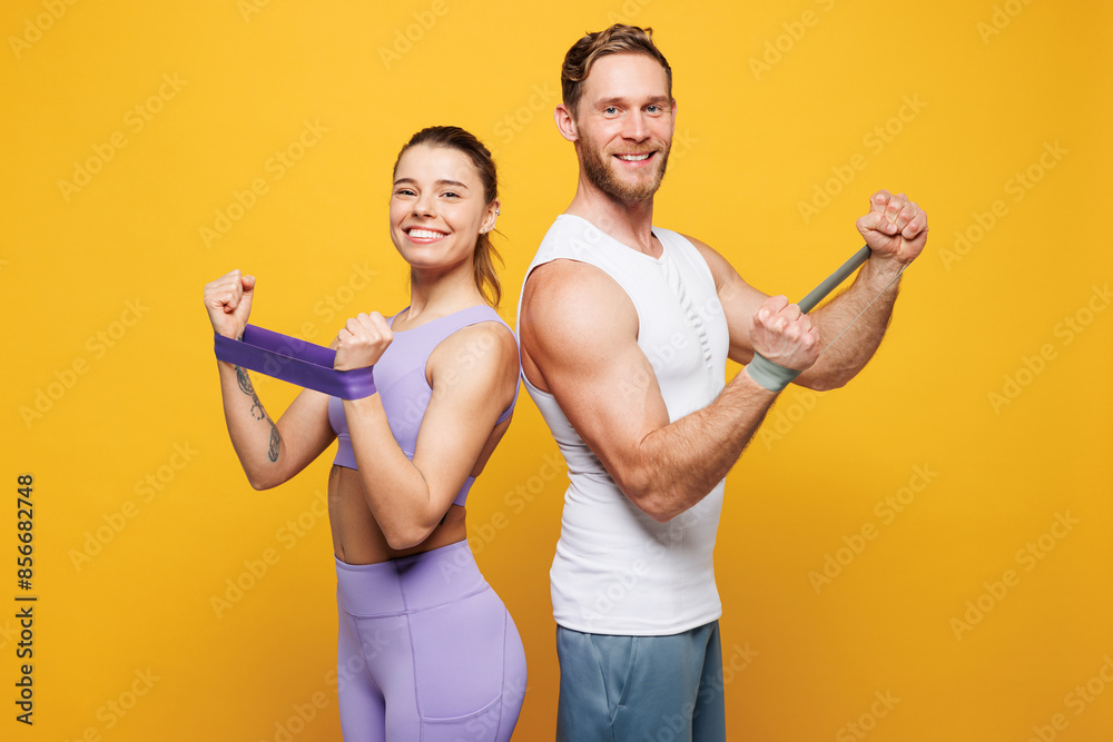 Young strong fitness trainer instructor sporty two man woman in blue clothes spend time in home gym hold in hand use elastic rubber band isolated on plain yellow background Workout sport fit concept Young strong fitness trainer instructor sporty two man woman in blue clothes spend time in home gym hold in hand use elastic rubber band isolated on plain yellow background Workout sport fit concept