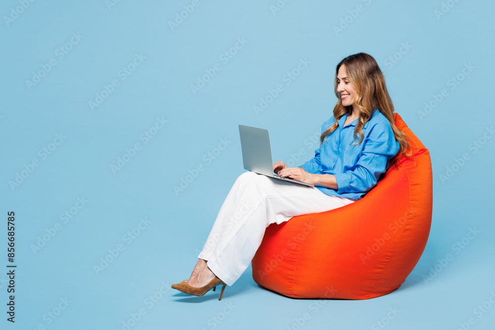 © ViDi Studio - Full body young smart IT woman she wear shirt casual clothes sit in bag chair hold use work on laptop pc computer isolated on plain pastel light blue cyan background studio portrait Lifestyle concept