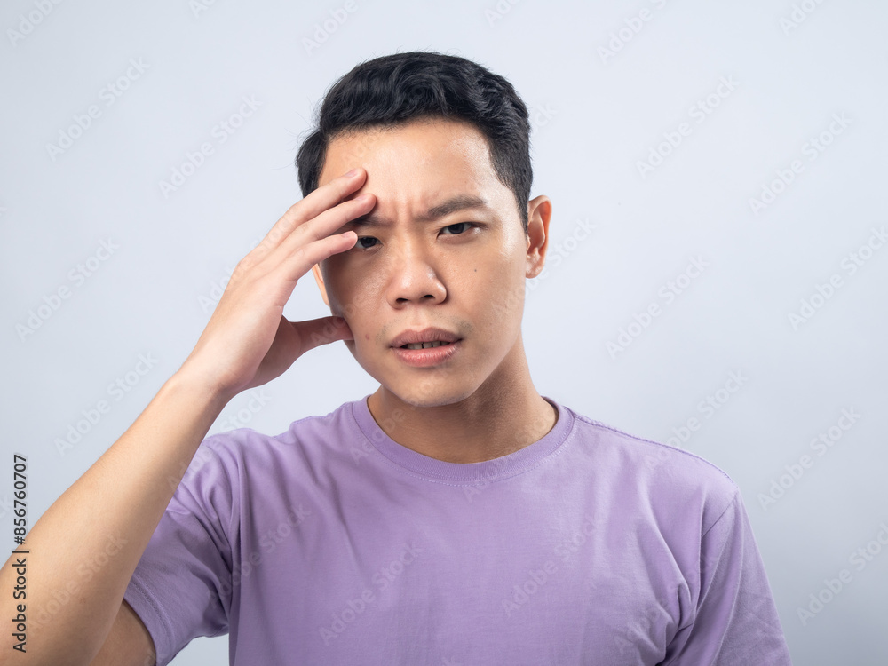 Asian man in a light purple shirt with a worried expression, touching his forehead in a gesture of concern, standing against a plain light grey background Asian man in a light purple shirt with a worried expression, touching his forehead in a gesture of concern, standing against a plain light grey background