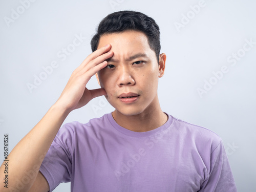 Asian man in a light purple shirt with a worried expression, touching his forehead in a gesture of concern, standing against a plain light grey background