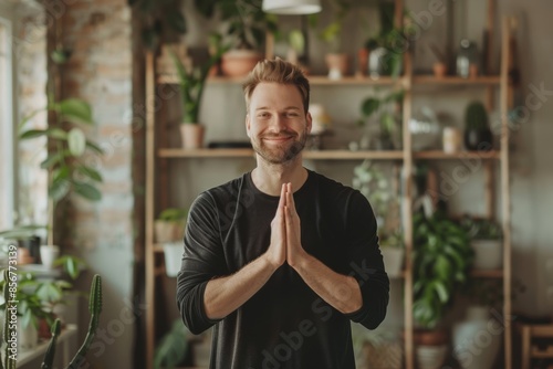 Portrait of a blissful caucasian man in his 20s joining palms in a gesture of gratitude in front of scandinavian-style interior background