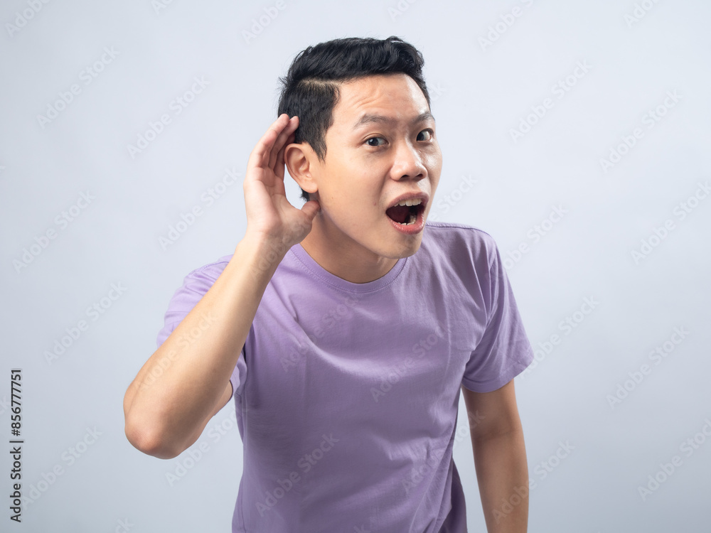 Young Asian man in a lavender t-shirt cupping his ear and looking surprised as if trying to hear something better. Studio shot on a plain background, highlighting his curious expression Young Asian man in a lavender t-shirt cupping his ear and looking surprised as if trying to hear something better. Studio shot on a plain background, highlighting his curious expression