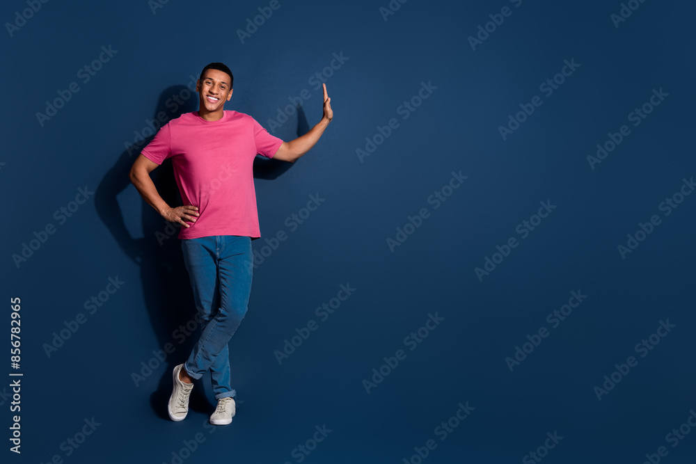 Full body length photo of young guy in pink t shirt and denim jeans confident standing near big offer advert isolated on dark blue color background Full body length photo of young guy in pink t shirt and denim jeans confident standing near big offer advert isolated on dark blue color background