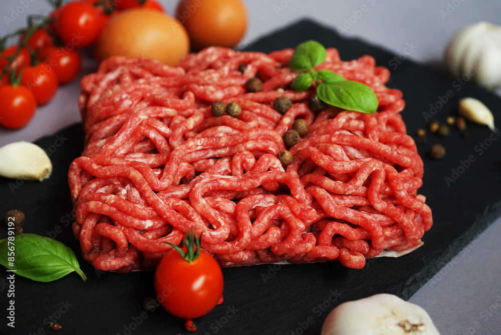 Fresh minced meat on a black slate tray next to cherry tomatoes, basil, eggs and spices on a gray background Fresh minced meat on a black slate tray next to cherry tomatoes, basil, eggs and spices on a gray background