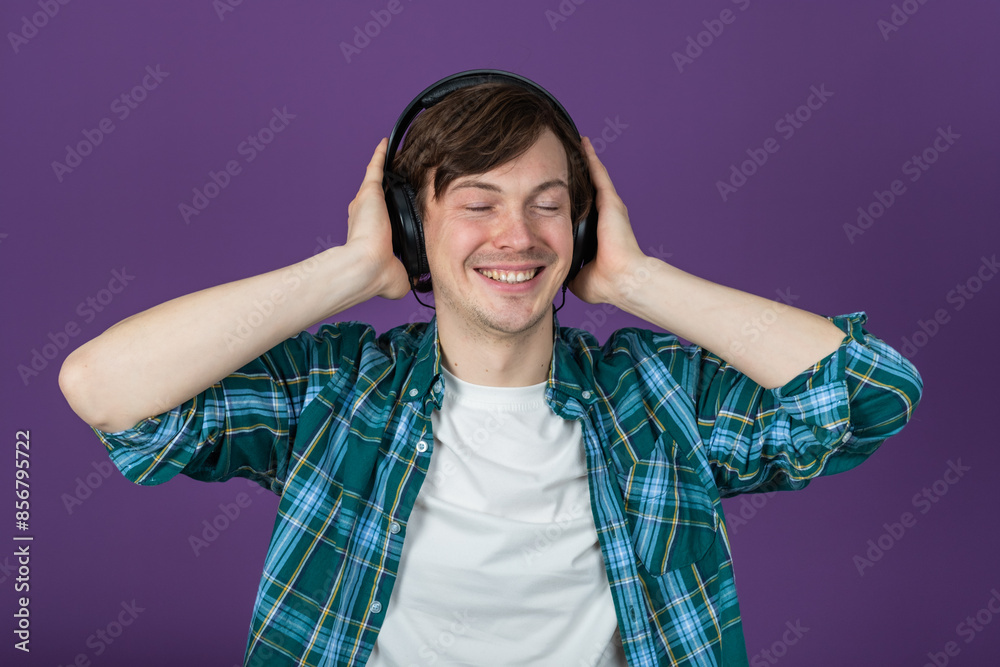 Young man in a green plaid shirt smiling and enjoying music with headphones on a purple background. Guy listens to his favorite tracks, pressing the headphones to his ears with his hands. Young man in a green plaid shirt smiling and enjoying music with headphones on a purple background. Guy listens to his favorite tracks, pressing the headphones to his ears with his hands.