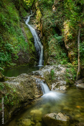 A serene waterfall cascades down mossy rocks in a vibrant forest near Klaus an der Pyrhnbahn, Austria. The sunlight filters through the canopy, illuminating the clear pool of water below.