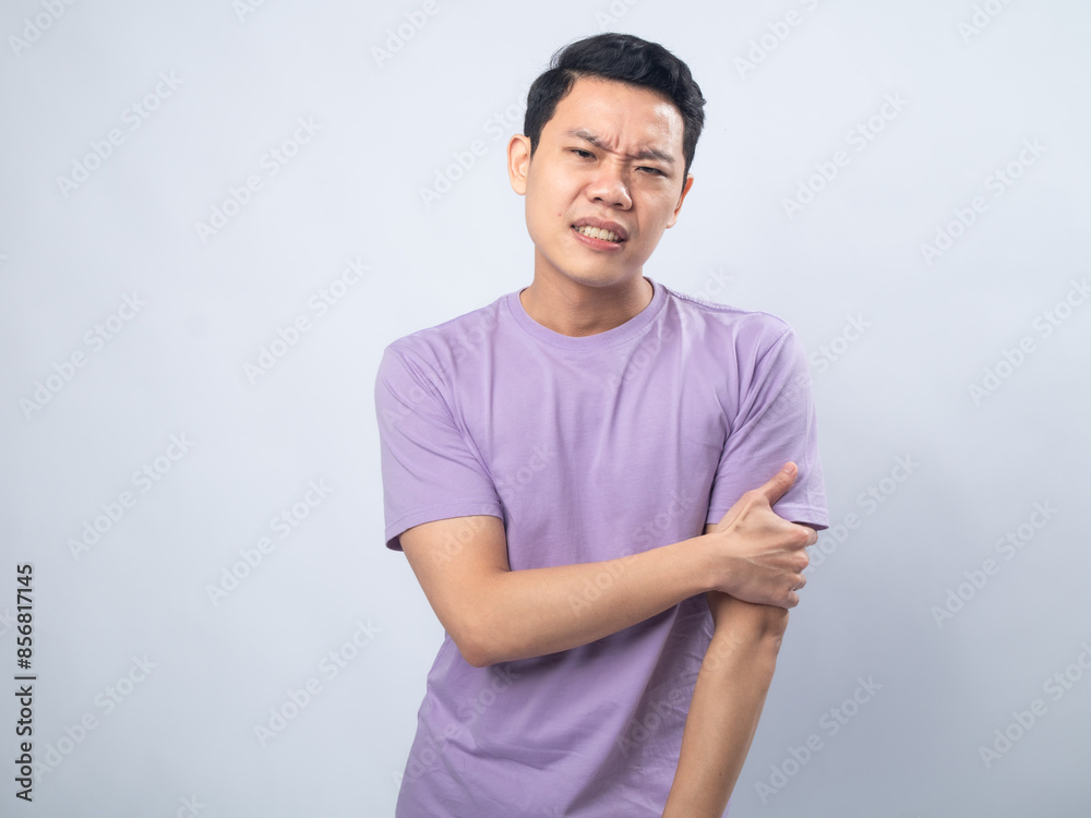 Obraz premium Young Asian man in a light purple shirt, wincing in pain while holding his left arm, standing against a plain light grey background, conveying discomfort or injury.
