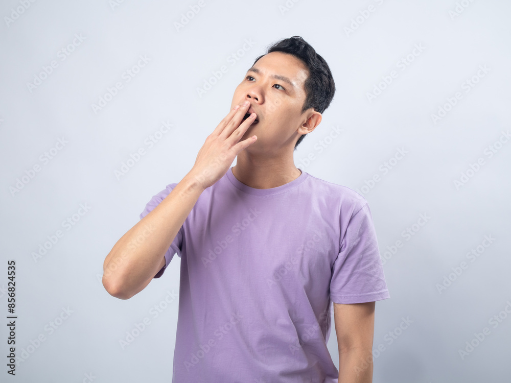 Young Asian man in a lavender t-shirt yawning and covering his mouth with his hand. Studio shot on a plain background, highlighting his sleepy expression and casual fashion. Young Asian man in a lavender t-shirt yawning and covering his mouth with his hand. Studio shot on a plain background, highlighting his sleepy expression and casual fashion.