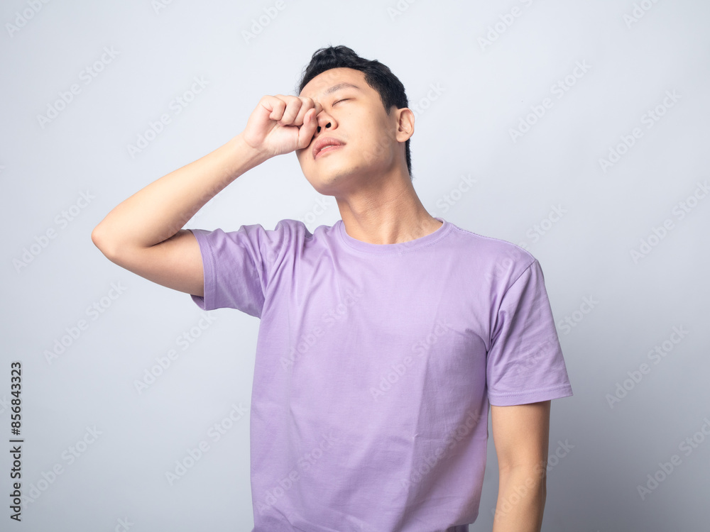 Young Asian man in a light purple shirt standing against a plain light grey background, rubbing his eyes with his left hand. The man's facial expression suggests fatigue or stress, Young Asian man in a light purple shirt standing against a plain light grey background, rubbing his eyes with his left hand. The man's facial expression suggests fatigue or stress,