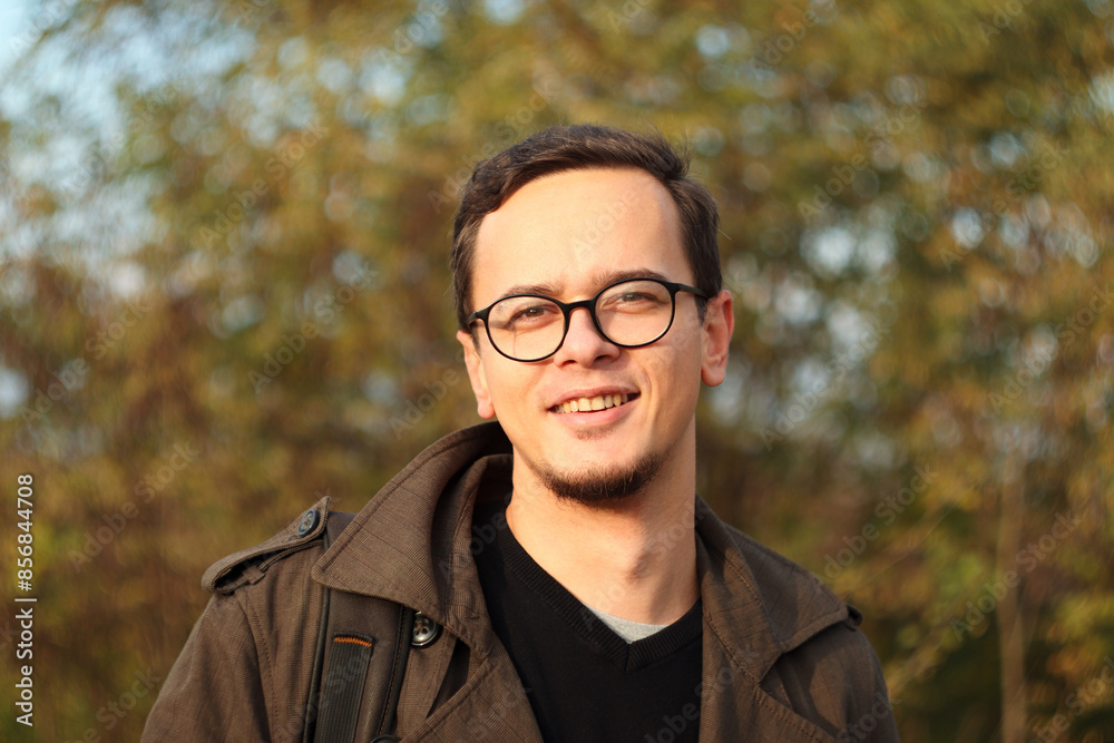 Cheerful young man with glasses, smiling in autumn sun. Happy male outdoors in sunny fall nature. Autumn man nature Cheerful young man with glasses, smiling in autumn sun. Happy male outdoors in sunny fall nature. Autumn man nature