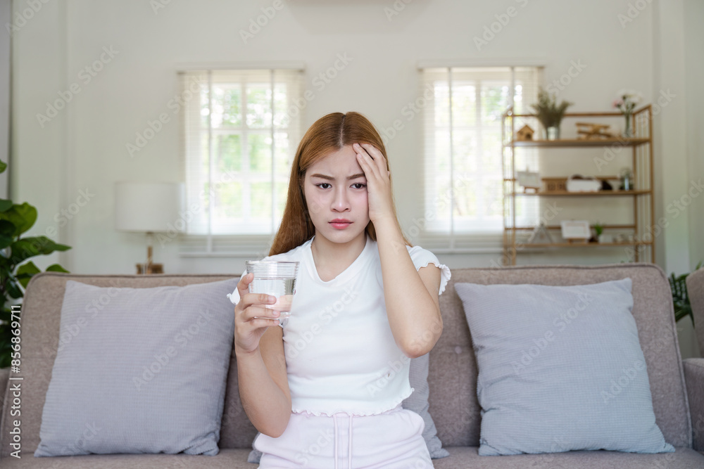 Young Woman Suffering from Headache Holding a Glass of Water While Sitting on a Sofa in a Bright Living Room Young Woman Suffering from Headache Holding a Glass of Water While Sitting on a Sofa in a Bright Living Room