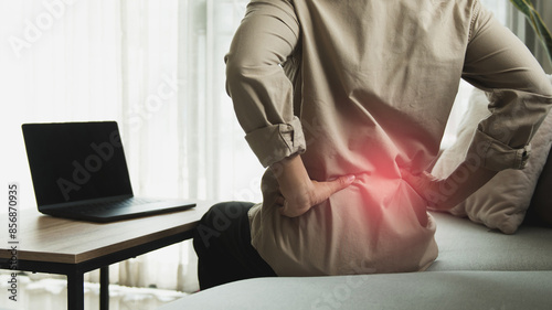 Working businessman sitting on the sofa in the living room at home He used both hands to press down on the lower back. He is suffering from back pain from sitting for a long time.