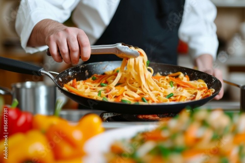 Close Up Photo Of Man’s Hands Serving Pasta With Fresh Vegetables An anonymous chef in apron picking up tagliatelle with serving tongs from the frying pan and serving it on the plate.