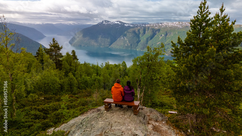 Konstfotografi A couple sits on a bench overlooking a Norwegian fjord, surrounded by lush greenery and towering mountains