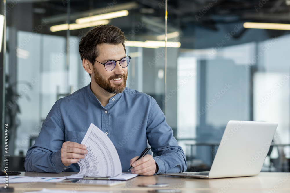 © Tetiana - A smiling young man sits in the office at a table in front of a laptop, makes notes in documents, works with data