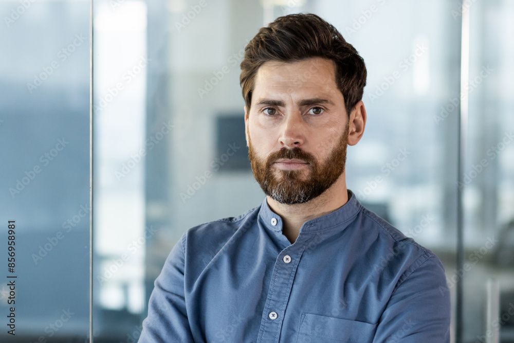 Naklejka premium Close-up portrait of a young serious businessman in a blue shirt standing inside the office and looking confidently at the camera