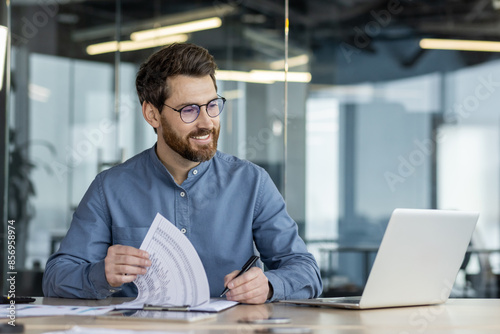 Fotografie A smiling young man sits in the office at a table in front of a laptop, makes no