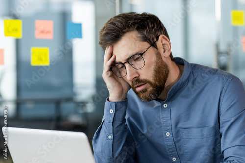 Tired and worried young businessman man sitting at workplace in front of laptop and upset with hand on head. Close-up photo