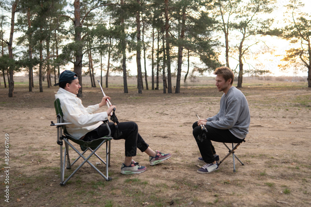 Two men sit on folding chairs in a forest clearing during a sunny afternoon. They are enjoying a picnic in the shade of the trees, with a peaceful and serene atmosphere Two men sit on folding chairs in a forest clearing during a sunny afternoon. They are enjoying a picnic in the shade of the trees, with a peaceful and serene atmosphere