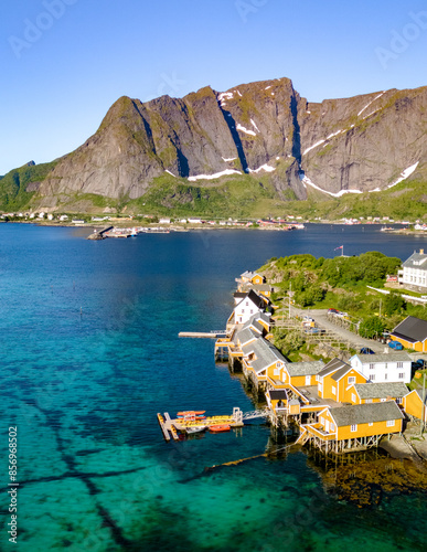 Aerial view of a small Norwegian village with colorful houses on stilts along the turquoise waters of a fjord. Mountains rise in the background, providing a picturesque backdrop. Sakrisoy, Lofoten photo