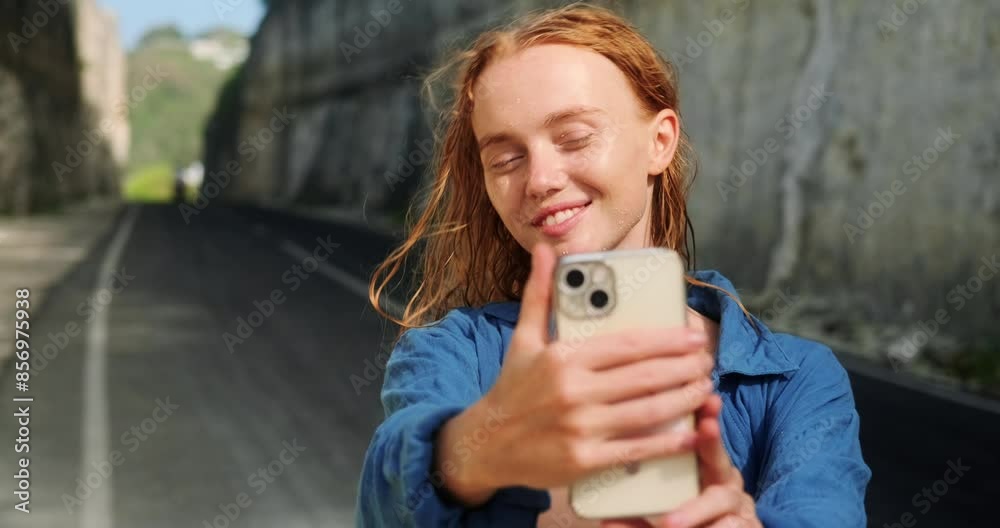 A young red haired woman with freckles takes a selfie on her phone, she looks at the camera and smiles. A travel blogger takes a photo against the backdrop of a landmark, a road between two rocks. A young red haired woman with freckles takes a selfie on her phone, she looks at the camera and smiles. A travel blogger takes a photo against the backdrop of a landmark, a road between two rocks.