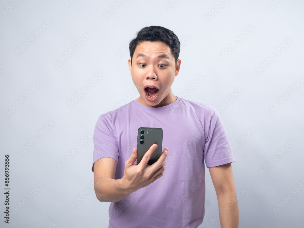 Young Asian man in a lavender t-shirt looking surprised and excited while waving at his smartphone. Studio shot on a plain background, highlighting his enthusiastic expression and casual fashion.