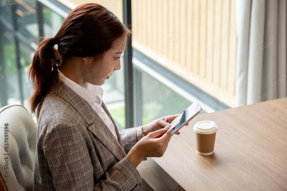 A charming, positive Asian businesswoman in a trendy suit sits in a coffee shop, checking messaged or replying chat on her smartphone. people and wireless technology concepts A charming, positive Asian businesswoman in a trendy suit sits in a coffee shop, checking messaged or replying chat on her smartphone. people and wireless technology concepts