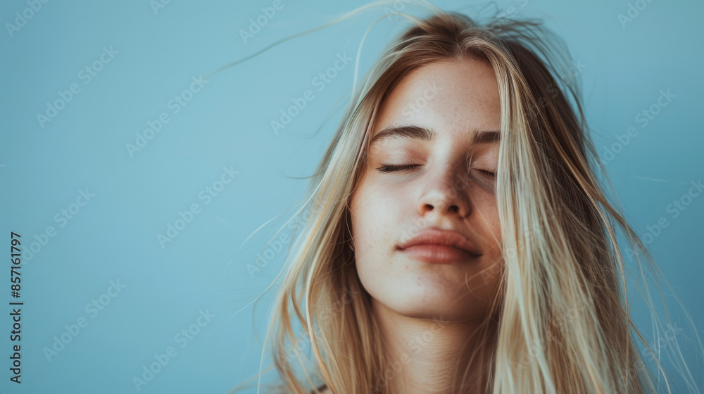 A young woman with closed eyes enjoys a peaceful moment, hair gently tousled against a soft blue background. A young woman with closed eyes enjoys a peaceful moment, hair gently tousled against a soft blue background.