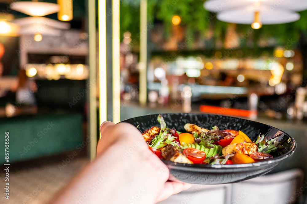Fototapeta premium Close-up of a hand holding a colorful salad on a black plate in a restaurant setting