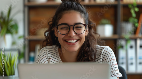 Cheerful young woman wearing glasses, smiling and working on a laptop in a cozy home office