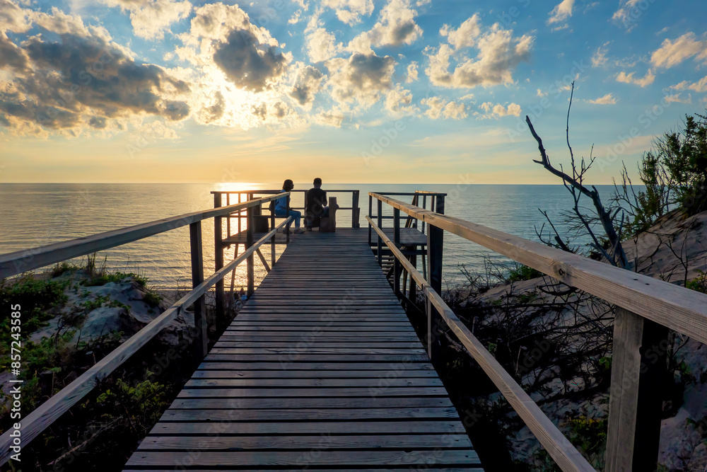 Naklejka premium A perspective of a wooden boardwalk across a dune. In the distance there are dark silhouettes of people sitting, admiring a beautiful sunset over the sea.
