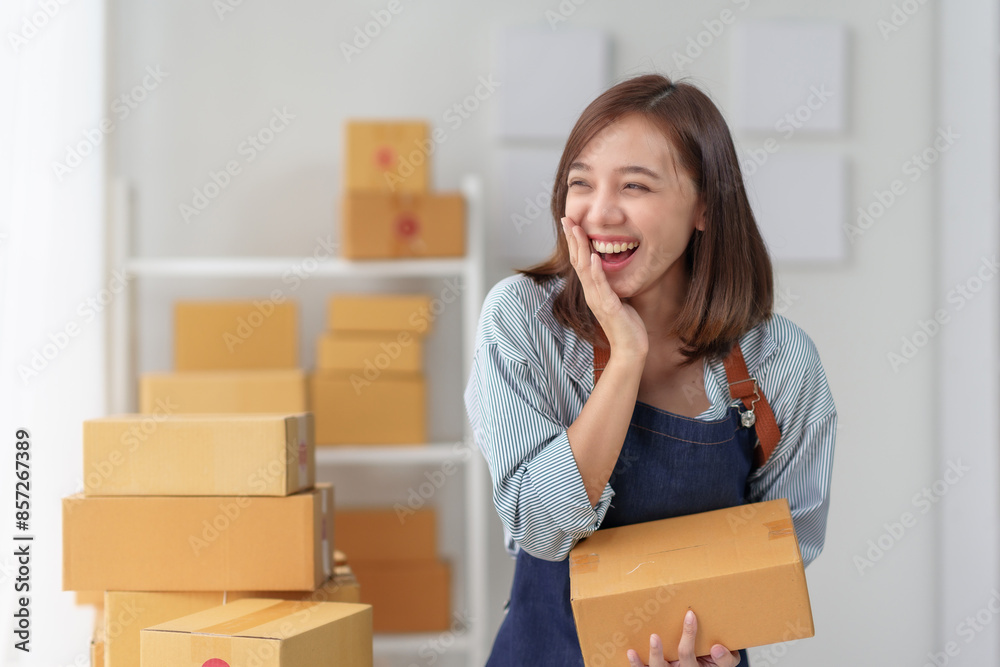 A cheerful woman holding a package in a workspace filled with boxes, representing online business, shipping, and e-commerce success. A cheerful woman holding a package in a workspace filled with boxes, representing online business, shipping, and e-commerce success.