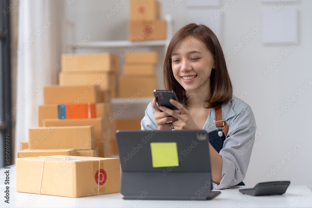 Young entrepreneur smiling while handling online business orders with a smartphone and laptop, surrounded by packages in a home office. Young entrepreneur smiling while handling online business orders with a smartphone and laptop, surrounded by packages in a home office.