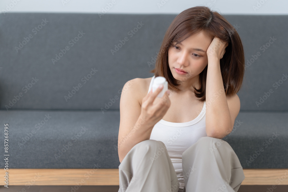 Woman looking at a small white object while sitting on the floor with a worried expression. Gray couch in the background. Woman looking at a small white object while sitting on the floor with a worried expression. Gray couch in the background.