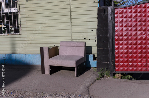 a broken upholstered armchair on the street against the background of a yellow wall with a blue foundation and a red gate
