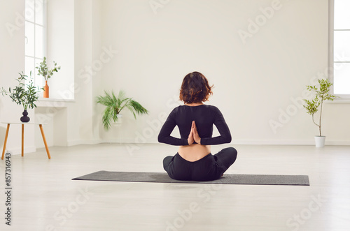 Yogi fit woman in sportswear doing yoga exercises for spine, shoulders, wrists, making Namaste gesture behind the back, sitting on mat, rear view in studio, home white background interior design