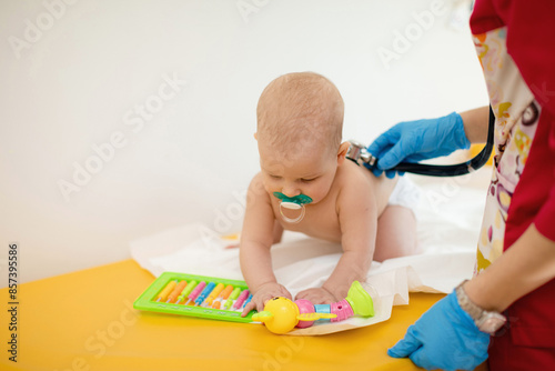 A pediatrician examining a baby using a stethoscope. A baby with pacifier is playing with a toy while being examined by a doctor.